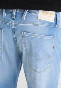 Šviesiai mėlynai deniminius džinsus su šiek tiek dėvėtu paviršiumi. Yra standartiniai užpakaliniai kišenės ir logotipo etiketas "REPLAY". Geltoni siuvimo detalės.