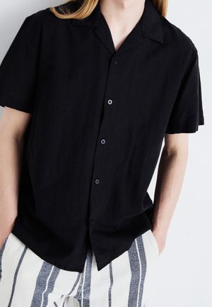 Personne portant une chemise noire à manches courtes boutonnée et un pantalon blanc avec des rayures noires verticales, debout les mains dans les poches.