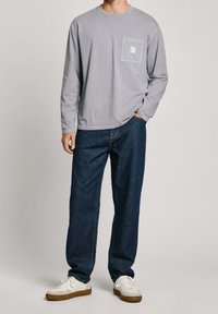 Hellgraues Langarmshirt mit einer Brusttasche, kombiniert mit dunkelblauen Jeans und weißen Sneakers mit beigen Sohlen. Lässiger Schnitt.