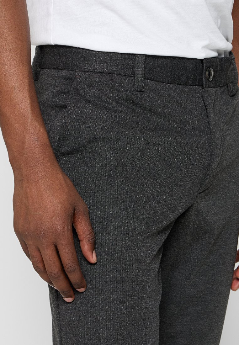 Ciemnoszare spodnie z fakturowanej tkaniny, z bocznymi kieszeniami, zapięciem na guzik i gładkim paskiem w talii. Zbliżenie.