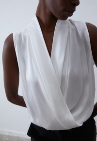 Ärmlös vit blus med draperad design, mjukt tyg och djup V-ringning. Slät struktur med diskret lyster. Svarta detaljer vid nederkant.
