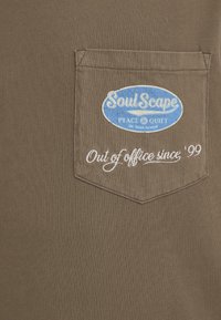 T-shirt en coton marron avec une poche poitrine. Présente une étiquette ovale bleue portant l'inscription « SoulScape » et un texte brodé blanc « Hors du bureau depuis 99 ».