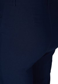 Marinblå byxor i slätt tyg, med en platt front och rena sömmar. Förvaringsdetalj synlig på vänster sida.