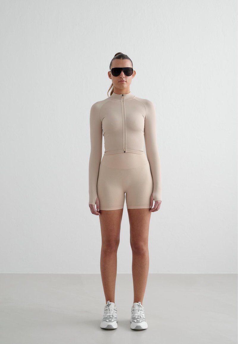 Ensemble de sport ajusté beige avec manches longues et shorts, fabriqué en matériau extensible lisse. Comprend une fermeture éclair sur le devant et des chaussures assorties.