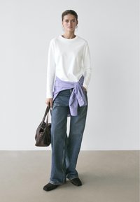 Woman standing wearing white long-sleeve shirt, purple shirt tied at waist, blue wide-leg jeans, black square-toe shoes, holding brown handbag.