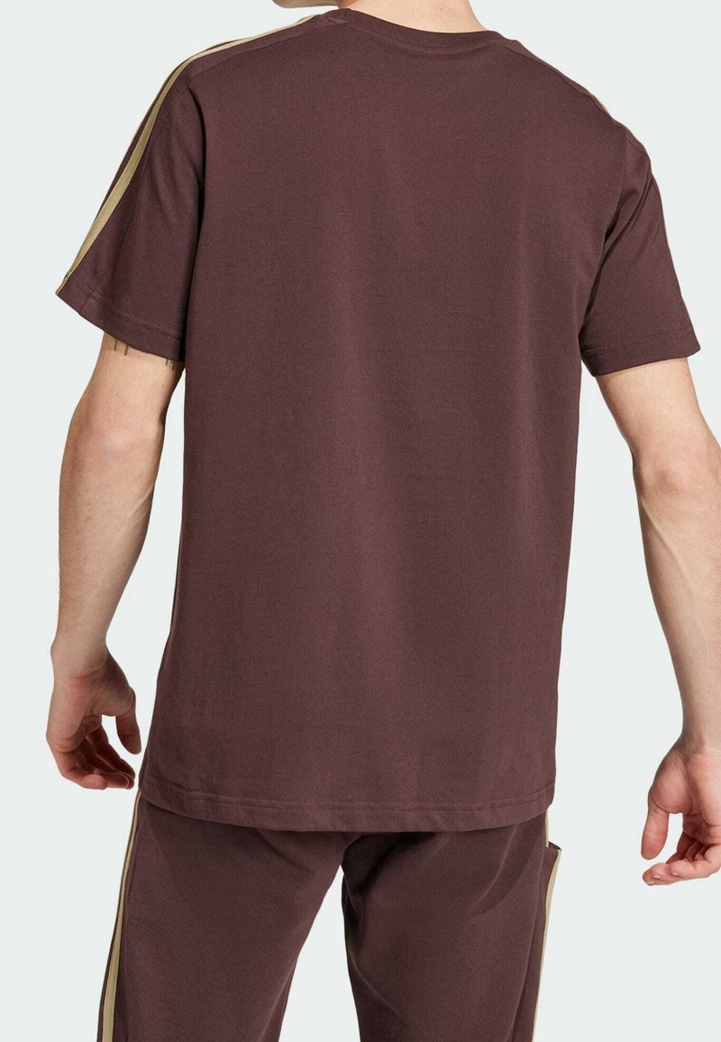 Hnědé bavlněné tričko s krátkým rukávem, které má zlaté pruhy podél ramen. Tkanina je hladká a tričko má volný střih.