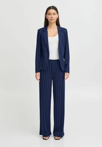 Costume bleu marine à rayures avec une veste ajustée et un pantalon large, fabriqué dans un tissu lisse avec un léger éclat. Haut blanc en dessous.