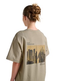 Person med fletning iført beige T-shirt med tryk på ryggen af bybygninger og et bjerg med teksten "HALO 1968 1972 1981" og "OFF DUTY GEAR."