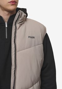 Svijetlosmeđi prsluk s punjenjem i patentnim zatvaračem, s crnim PGDR logotipom i crnom duksericom ispod, prikazuje kviltanu teksturu.