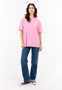 Overmaatse roze katoenen t-shirt met een grafische borstzak, gecombineerd met blauwe denim jeans en zwarte lage sneakers. Eenvoudig en casual ontwerp.