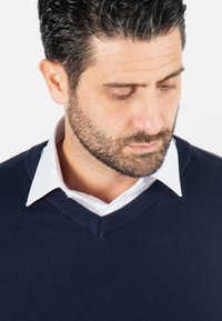 Pull en maille bleu marine avec un col en V, porté sur une chemise blanche à col. Le tissu semble lisse et léger, avec un fin côtelage sur les bords.