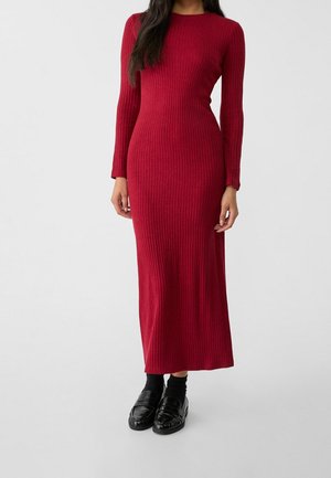 Kvinde iført en langærmet, ribstrikket, ankellang rød kjole kombineret med sorte sokker og skinnende sorte loafers, stående foran en ensfarvet baggrund.