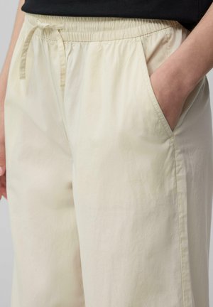 Jasno beżowe spodnie typu wide-leg wykonane z miękkiego materiału, z elastycznym paskiem w talii oraz sznurkiem i kieszeniami po bokach. Gładka struktura.