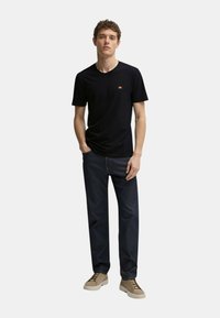 T-shirt nera a maniche corte con un piccolo logo arancione, abbinata a jeans blu scuro e sneakers beige chiaro con lacci marroni.