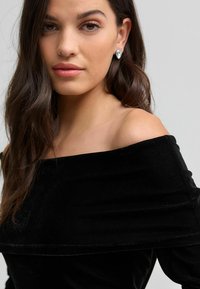 Svart avoff shoulder-topp i mjuk sammet med en slät yta och minimala sömmar. Modellen har långt, vågigt hår och enkla örhängen.