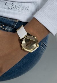 Montre dorée et blanche avec un cadran rond, design minimaliste et bracelet en caoutchouc. Affiche un marqueur d'heure triangulaire et un logo.