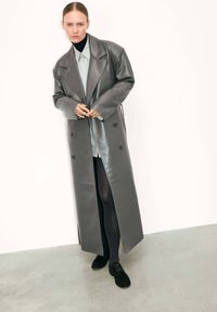 Femme portant un long manteau en cuir gris à double boutonnage, une chemise claire, des collants noirs et des chaussures noires, se tenant sur un sol gris contre un mur blanc.