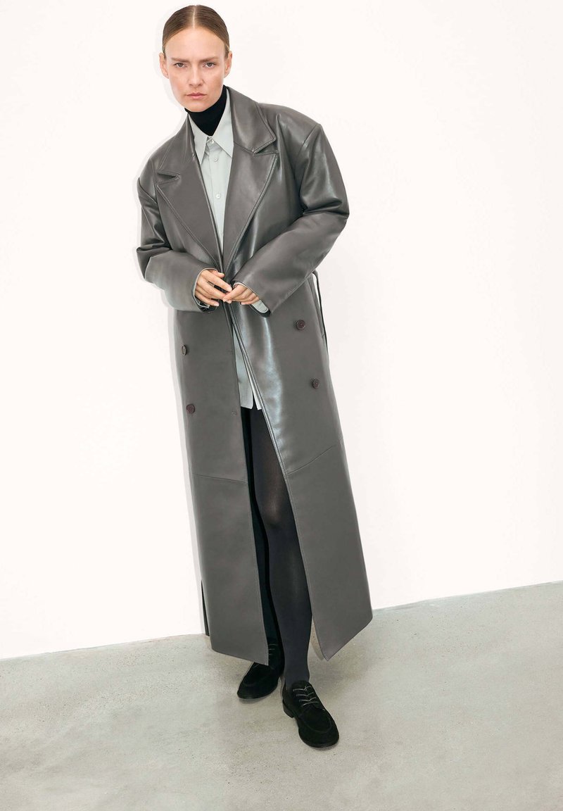 Femme portant un long manteau en cuir gris à double boutonnage, une chemise claire, des collants noirs et des chaussures noires, se tenant sur un sol gris contre un mur blanc.