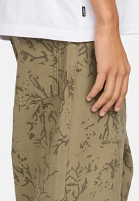 Un pantalon vert olive avec une surface texturée et un motif sombre subtil ressemblant à des branches. Comprend un détail de poche latérale.