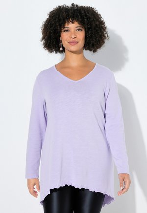 Hymyilevä henkilö, jolla on kiharat hiukset, värikkäät korvakorut, vaaleanvioletti pitkähihainen V-kaula-aukollinen paita, jossa on röyhelöreuna, ja mustat kiiltävät housut.