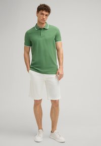 Grünes Poloshirt mit weißen Akzenten, kurzen Ärmeln und Logo. Kombiniert mit weißen Shorts und hellen Sneakern. Glatte Textur, lockere Passform.