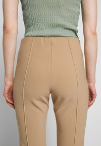 Beige tailored trousers with a smooth texture, high waist, and vertical seam details. Worn with a light green, ribbed top.