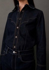 Chemise en denim foncé avec des coutures contrastées et des boutons en métal, associée à un jean en denim foncé taille haute sur un fond uni.