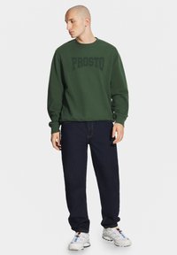 Zielony bawełniany sweter z prążkowanymi mankietami, z wypukłym logo "PROSTO" w ciemnozielonym kolorze. Połączony z ciemnoniebieskimi dżinsami i białymi trampkami.