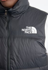 Black puffer vest with a high collar, featuring a front zipper and white embroidered logo. Smooth texture with a slight sheen.