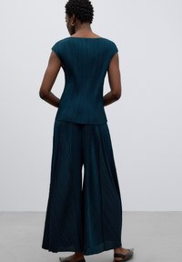 Une personne avec les cheveux bouclés courts se tient de dos, portant un haut plissé teal et un pantalon large, sur un fond gris clair.