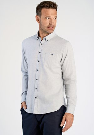 Homme portant une chemise à boutons gris clair avec une poche sur la poitrine et un pantalon sombre, regardant vers sa gauche sur un fond uni.