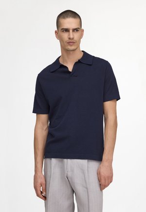 Mężczyzna ubrany w granatowy polo z krótkim rękawem oraz jasnoszare spodnie w cienkie prążki, stojący na jednolitym białym tle.