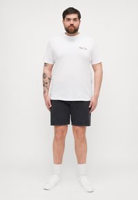 White cotton t-shirt with short sleeves and a small black logo. Worn with dark denim shorts and white sneakers. Solid colors, casual wear.