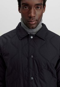 Gepolsterte schwarze Jacke mit spitzem Kragen, eleganten Knöpfen und einem strukturierten Wellenmuster. Trägt man über einem grauen Rollkragenpullover.