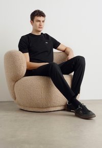 Beige plüss fotel lekerekített formával; egy fekete inget és nadrágot viselő személy kényelmesen ül, fekete cipő van rajta egy világos padlón.