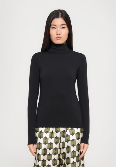 Femme aux longs cheveux noirs portant un col roulé noir ajusté et un pantalon ample avec un motif géométrique de cercles verts et blancs.