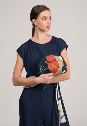 Mujer con vestido azul marino sosteniendo una pequeña bolsa con un patrón abstracto con formas rojas, beige y verde oscuro y texto en cursiva que dice "Anekke".