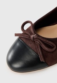 Brown suede flat with a black leather toe cap. Features a bow detail at the front and a soft interior lining.
