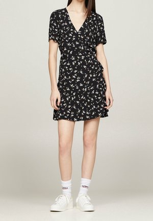 Femme portant une robe portefeuille noire à fleurs à manches courtes, avec des chaussettes blanches et des baskets blanches, se tenant devant un fond uni.