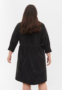 Robe noire, à mi-longueur, en tissu doux et texturé ; dotée de manches longues, d'une taille froncée et d'une coupe décontractée. La vue de dos montre une silhouette lisse.
