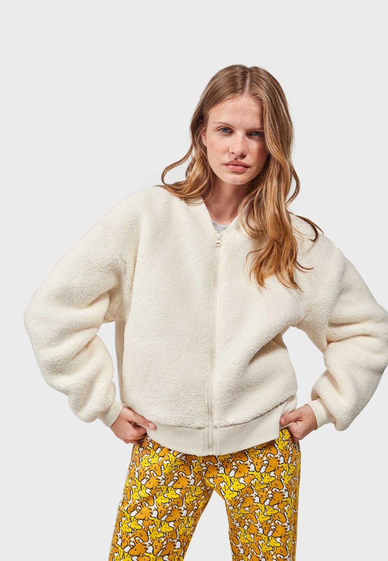 Witte, fluffy bomberjack met rits en ronde halslijn, afgewerkt met een ribbelsrand. Gecombineerd met levendige gele broek met een speelse print.