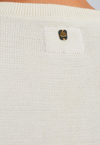 Maglione bianco a maglia con colletto a coste e un piccolo etichetta logo nero e oro cucita vicino al collo. Texture leggera.