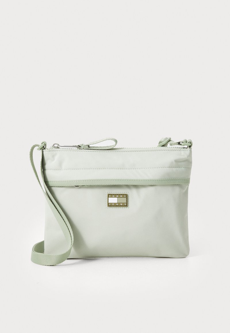 Bolsa transversal de nylon verde-claro com fecho em zíper, alça ajustável e detalhe em placa de logotipo em tom dourado. Textura suave e forma retangular.