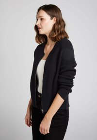 Gilet bleu marine foncé avec une fermeture éclair à l'avant, des poignets côtelés et un tricot texturé. Porté sur un haut blanc, avec une coupe décontractée.