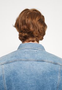 Ljusblå denimjacka sedd bakifrån, med en klassisk krage och synliga sömnadsdetaljer. Tyget har en mjuk, strukturerad yta.