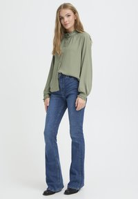 Svijetlo maslinasto zelena bluzica s naboranim dekolteom, dugim rukavima i gumbima sprijeda, uparena s plavim širokim trapericama i crnim čizmama do gležnja.