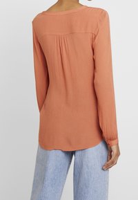 Blouse à manches longues de couleur rouille avec un tissu texturé, dos plissé, ourlet arrondi et poignets boutonnés, portée sur un jean bleu clair.