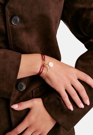 Verschränkte Frauenhände, die eine braune Wildlederjacke und ein bordeauxrotes Cord-Armband mit goldfarbenen Martini-Glas- und Kreis-Anhängern tragen.