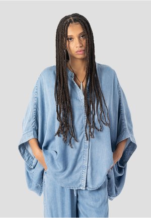 Kvinde med lange fletninger iført løs, lys denimskjorte og bukser med hænderne i lommerne, stående foran en ensfarvet grå baggrund.