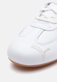 Baskets blanches avec un dessus en cuir lisse et une semelle en caoutchouc, présentant un logo Puma brodé en argent et des lacets blancs.